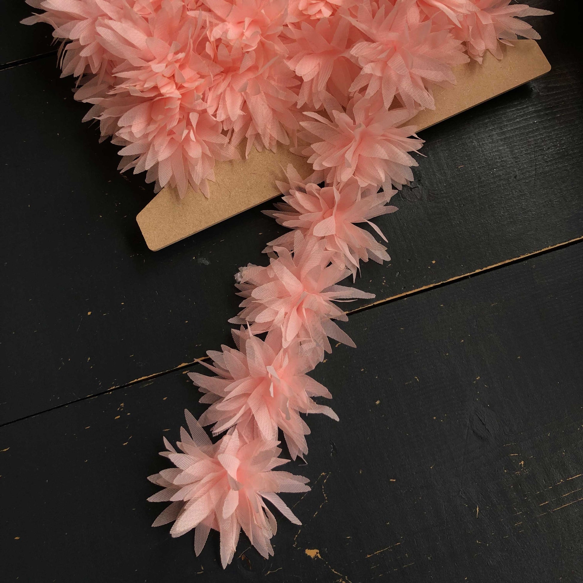 Dit speelse sierband is versierd met zachte, luchtige bloemetjes in een romantische zalmroze tint. De bloemen zijn gemaakt van een lichte, doorschijnende stof die subtiel meebeweegt